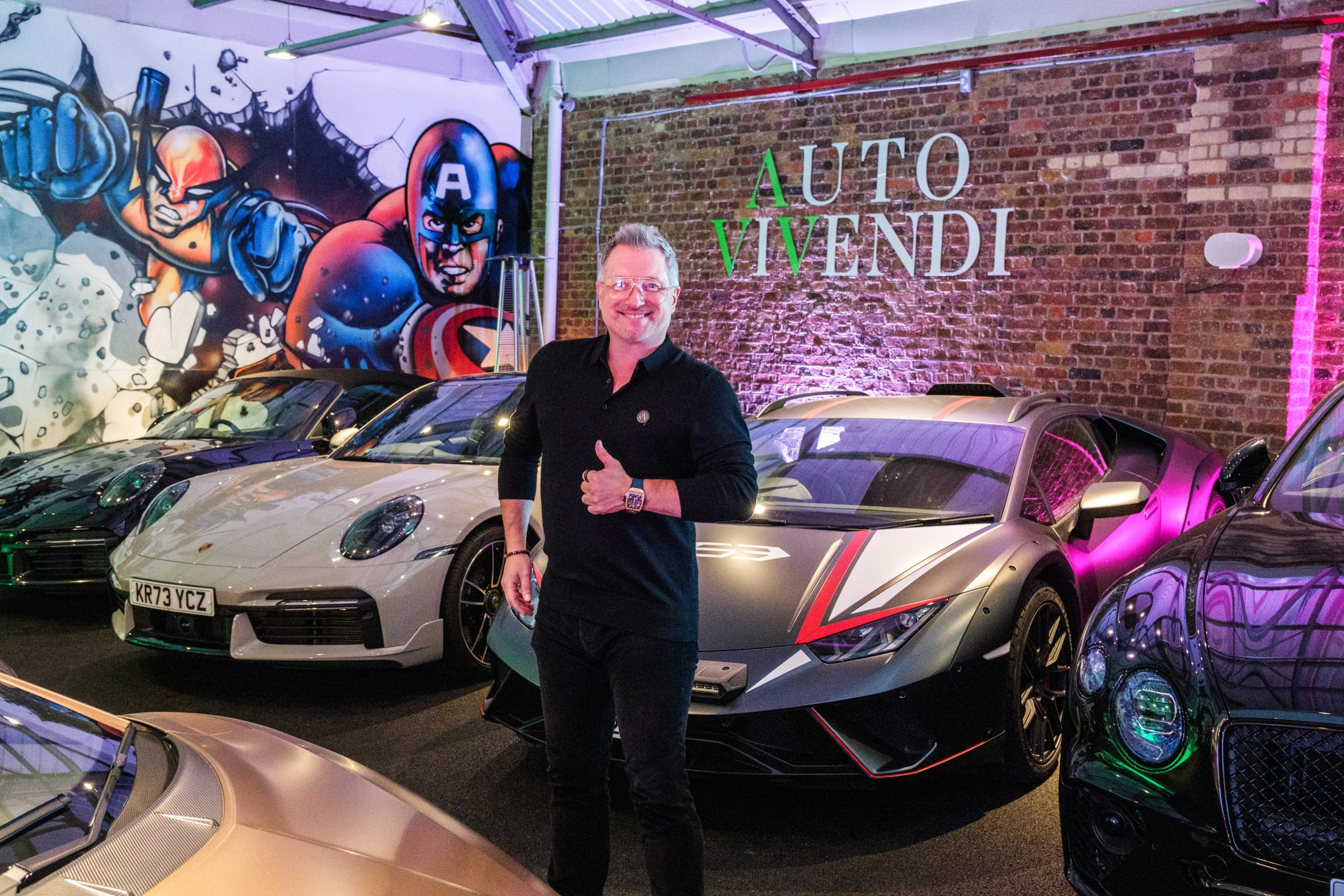 Smiling man in black shirt giving a thumbs up in a car showroom with luxury cars and a brick wall mural behind him