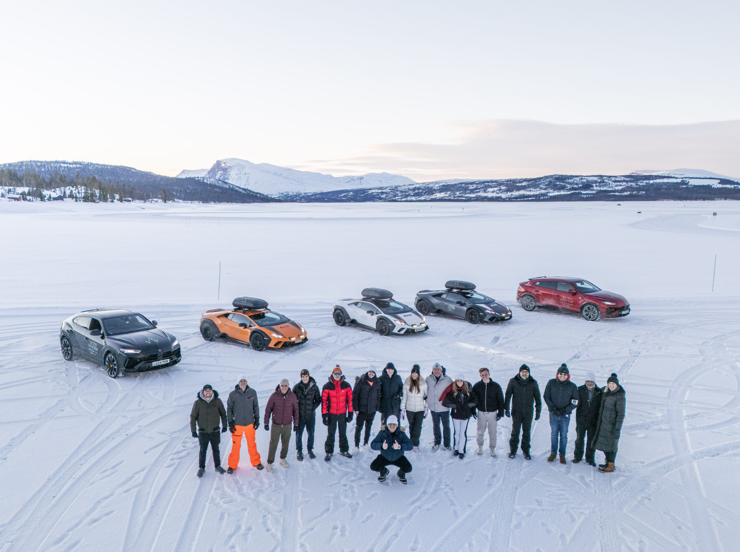 Group of people posing in a snowy field with several sports cars parked behind them and snow covered mountains in the distance